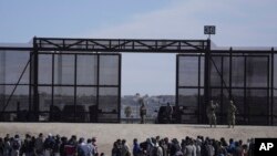 FILE - Migrants who crossed the border from Mexico into the US wait next to the US border wall where US Border Patrol agents stand guard, seen from Ciudad Juarez, Mexico, March 30, 2023. 